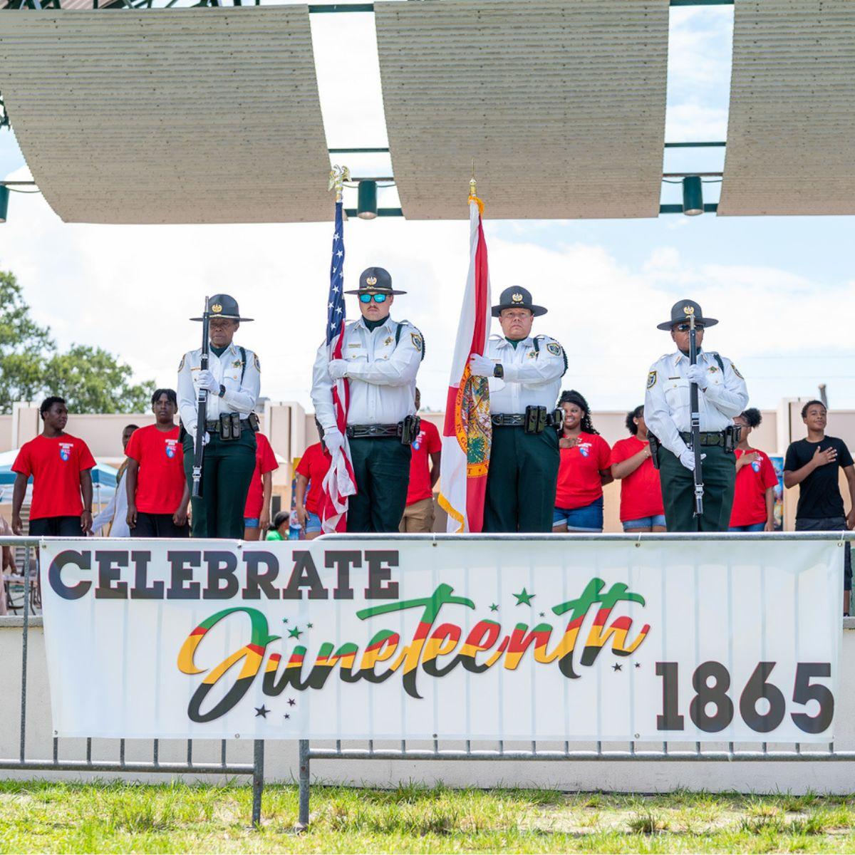 Honor guard stands on stage during a Juneteenth celebration ceremony, with the U.S. and Florida flags displayed above a “Celebrate Juneteenth 1865” banner while attendees stand in the background.
