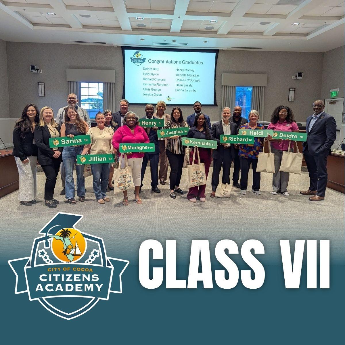 The graduating Citizens Academy class standing with elected officials and city staff inside of council chambers. Grads are holding street signs with their name on it. Also included is the citizens academy logo and the words "Class VII"