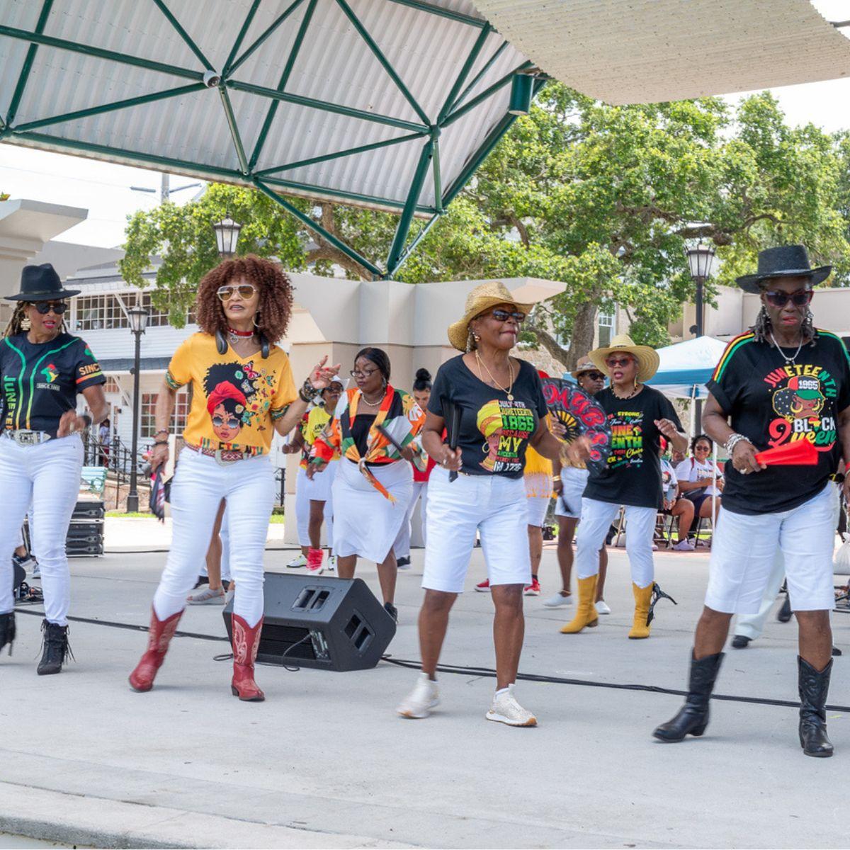 Group of women line dancing on an outdoor stage during a Juneteenth celebration, wearing colorful Juneteenth-themed shirts, white pants, cowboy boots, and hats while performing in front of a crowd.