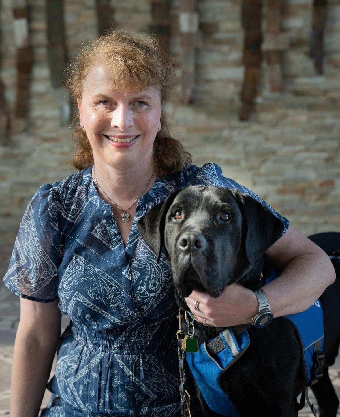 Kathy sits beside her guide dog Tate who is wearing a blue harness. She is dressed in a blue patterned blouse and has one arm around Tate. The pair are posed in front of a stone wall, and Tate looks directly at the camera while Kathy smiles warmly. 