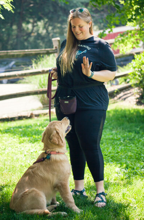 Catherine stands on green grass in an outdoor setting, holding a leash in one hand and raising the other hand in a training gesture. A dog sits attentively, looking up. Catherine wears a dark short-sleeve top, dark leggings, sandals, and a treat pouch. Behind them is a wooden fence and leafy trees, with sunlight creating bright patches on the grass.