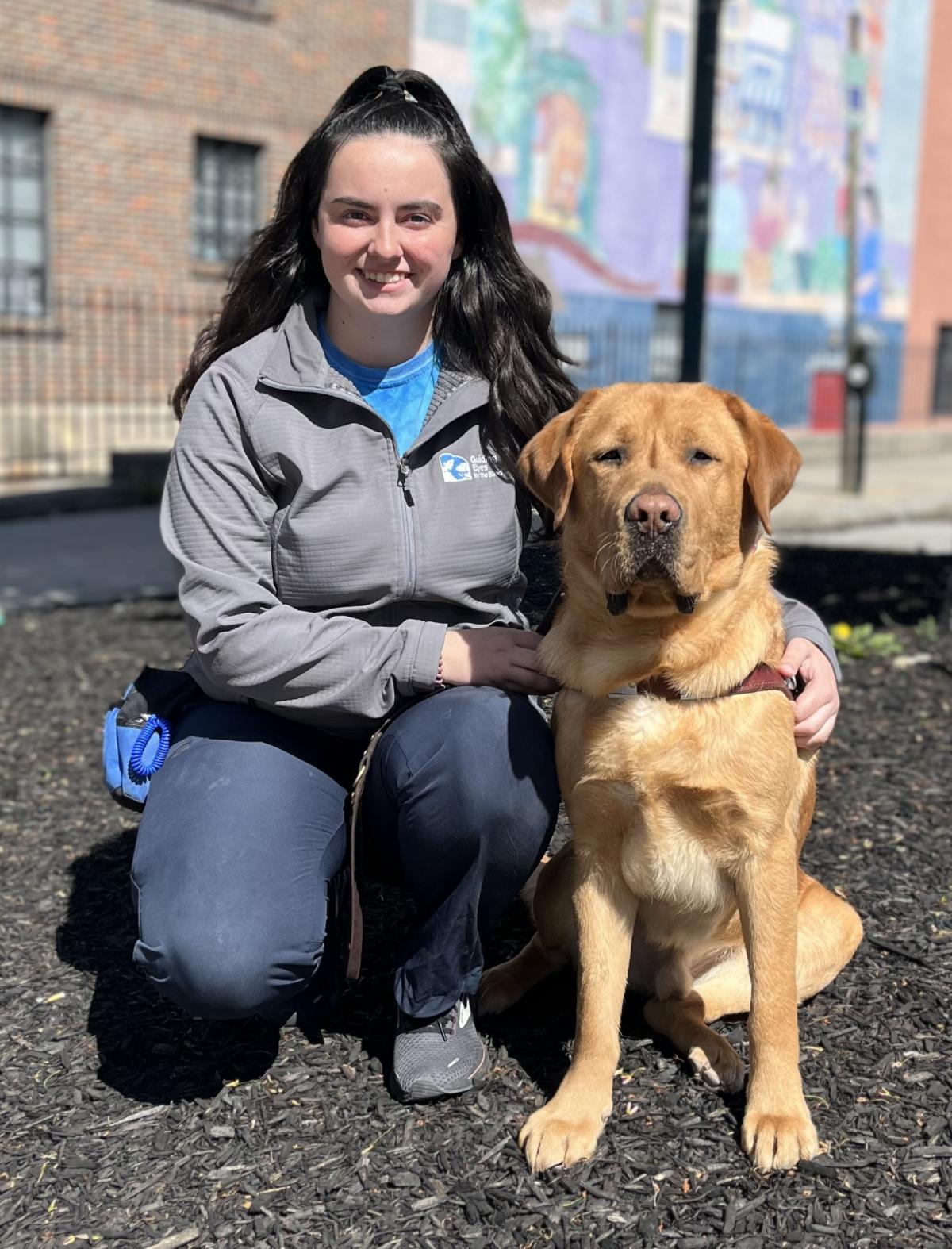 Erin kneels with her arm around medically released guide dog in training Siggy. Siggy is a large darker yellow lab with a big head, an adorable droopy face, and a small white patch on his chest. They are on some dark wood chips with a tree and some colorful/brick buildings in the background.