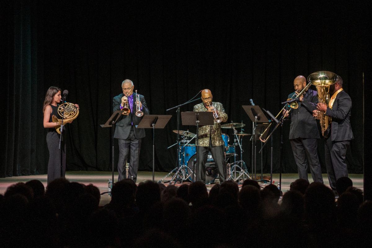 Five musicians play in a row on a stage with a black curtain