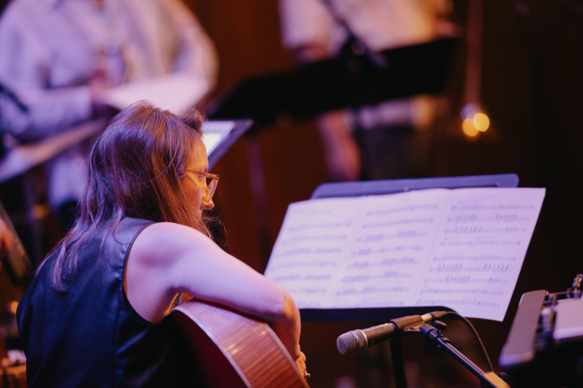A person with long hair and glasses is playing a guitar, seated in front of a music stand with sheet music. Other musicians are blurred in the background.