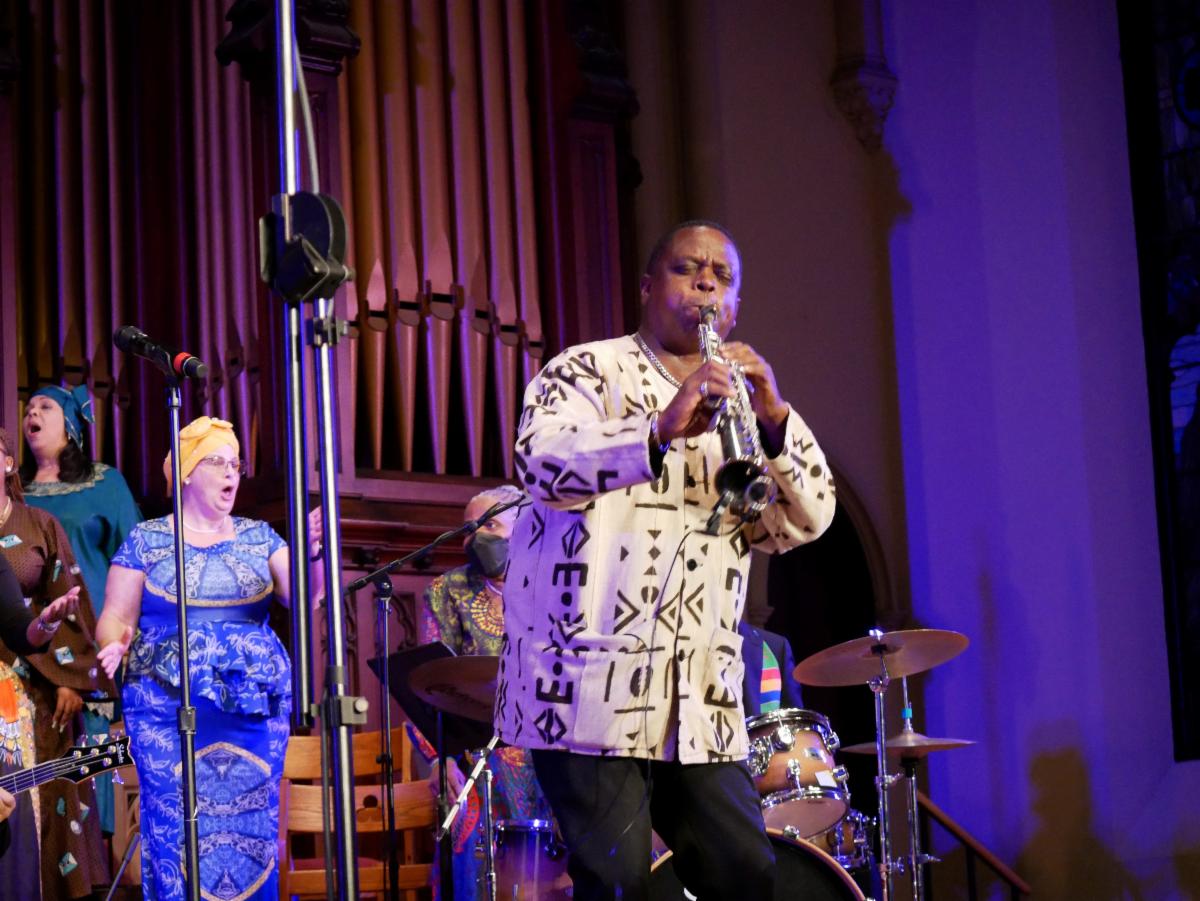 A musician wearing a white and black patterned shirt plays a woodwind instrument on a stage