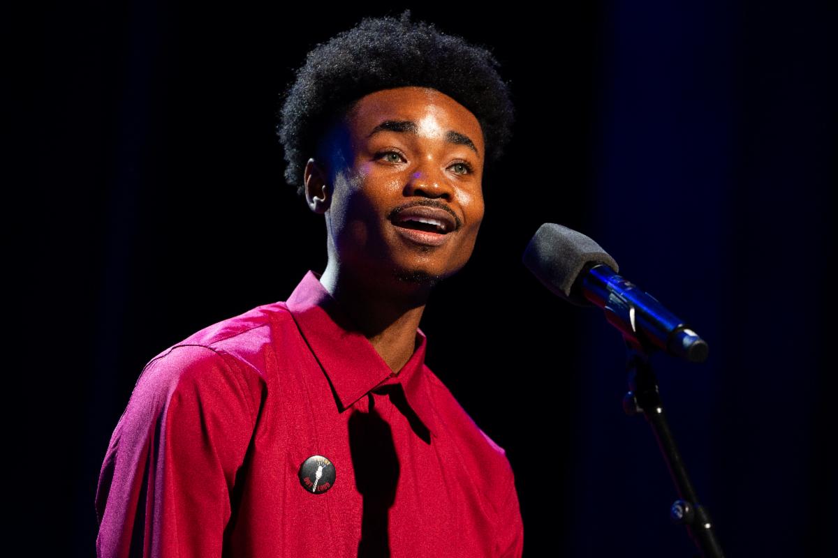 A teen boy in a red button down shirt recites poetry in front of a microphone