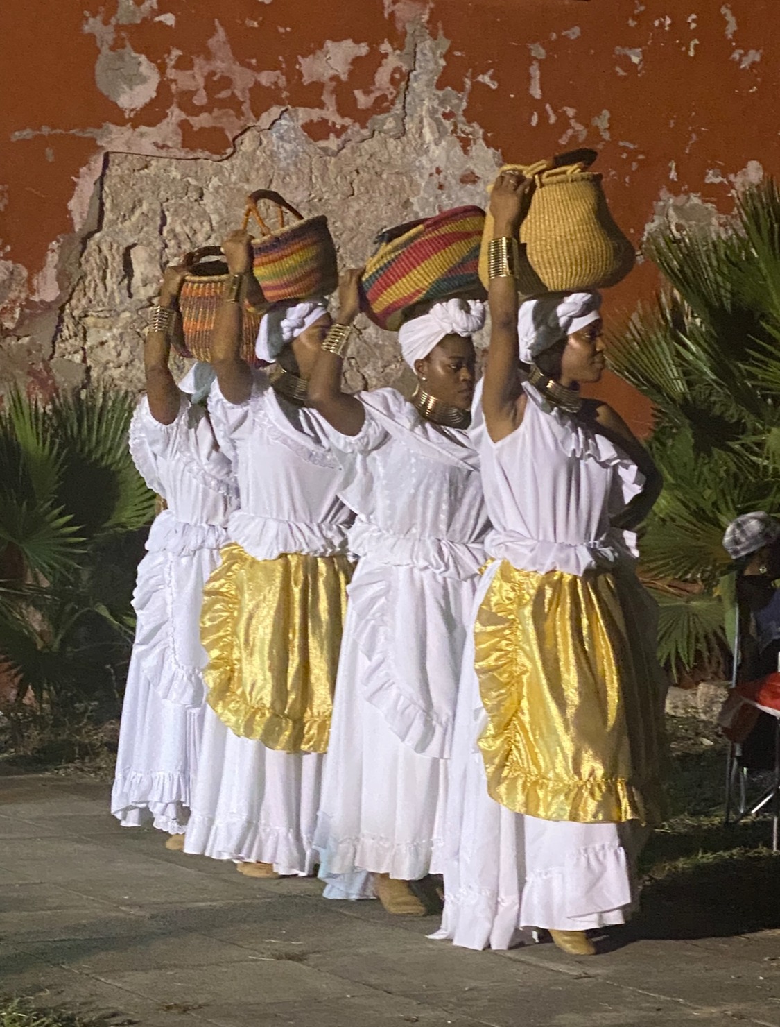 A row of women in a line wearing white dresses and holding baskets atop their heads