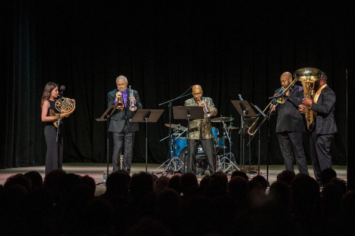 Five brass musicians stand on a black stage 