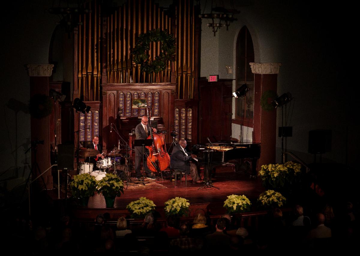 A music trio performs in front of a large organ on a wooden stage surrounded by plants