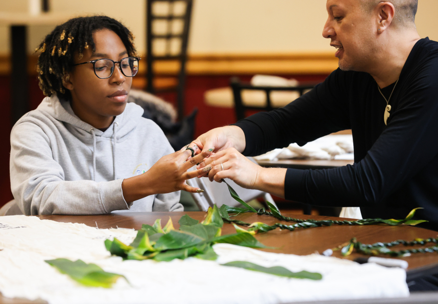 A teacher demonstrates lei making to a student at a table