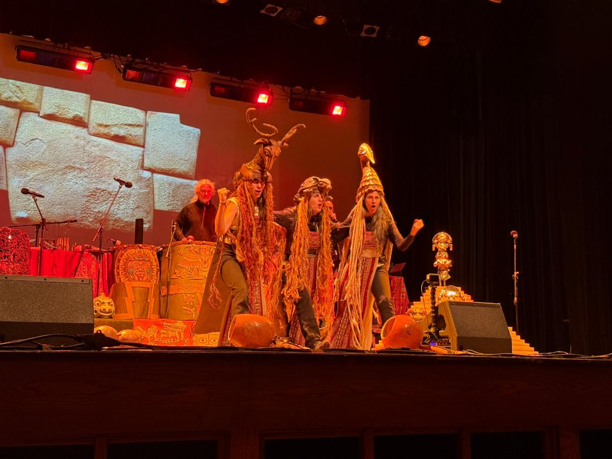 Members of the Orchestra of Indigenous Instruments and New Technologies perform at Pro-Art in Wise, VA, under red stage lighting. Performers wear elaborate costumes and headpieces, surrounded by decorated instruments. A stone wall backdrop is projected behind them.