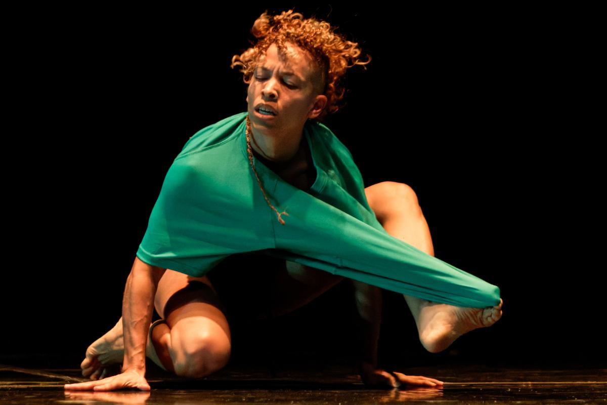 A dancer crouches on stage with a green shirt, stretching the shirt with their foot