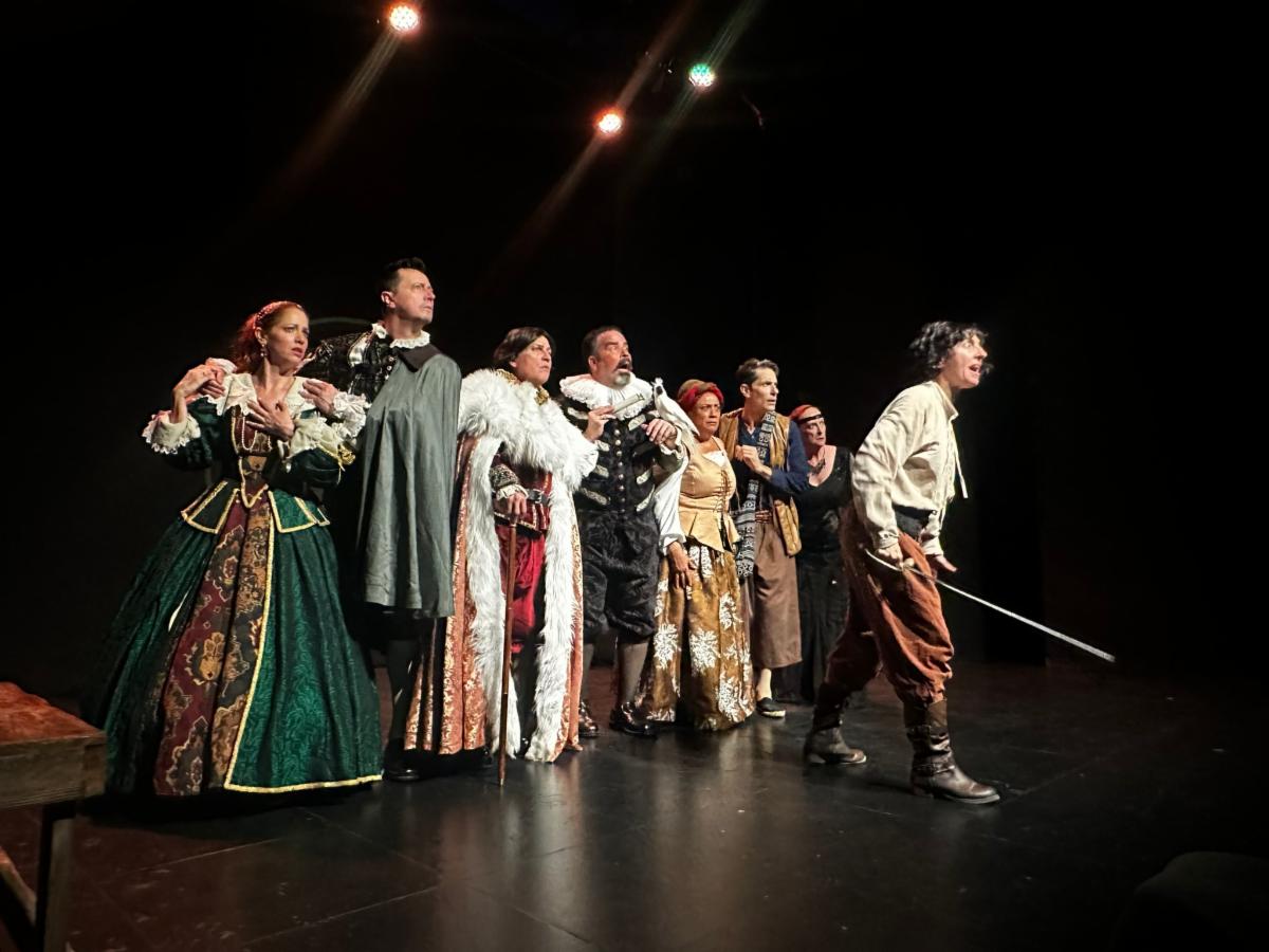 A group of performers in period costumes stands on stage, with one person holding a sword and looking forward.