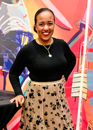 Nikki smiles in front of a red wall, smiling in a black long sleeved shirt and a brown and black patterned skirt