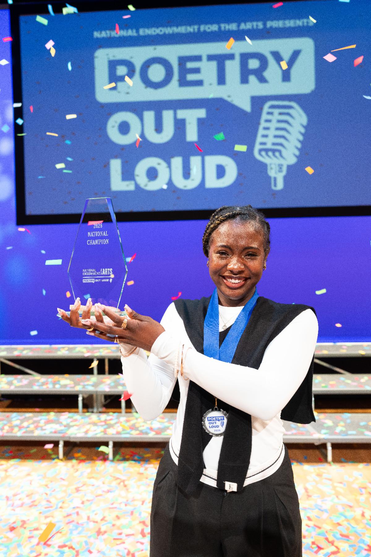A girl smiles holding a trophy in front of a blue Poetry Out Loud banner