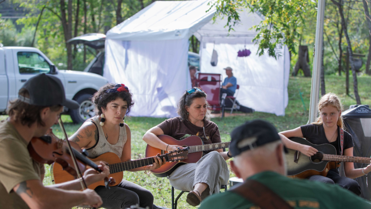 A group of musicians play stringed instruments outside in a circle
