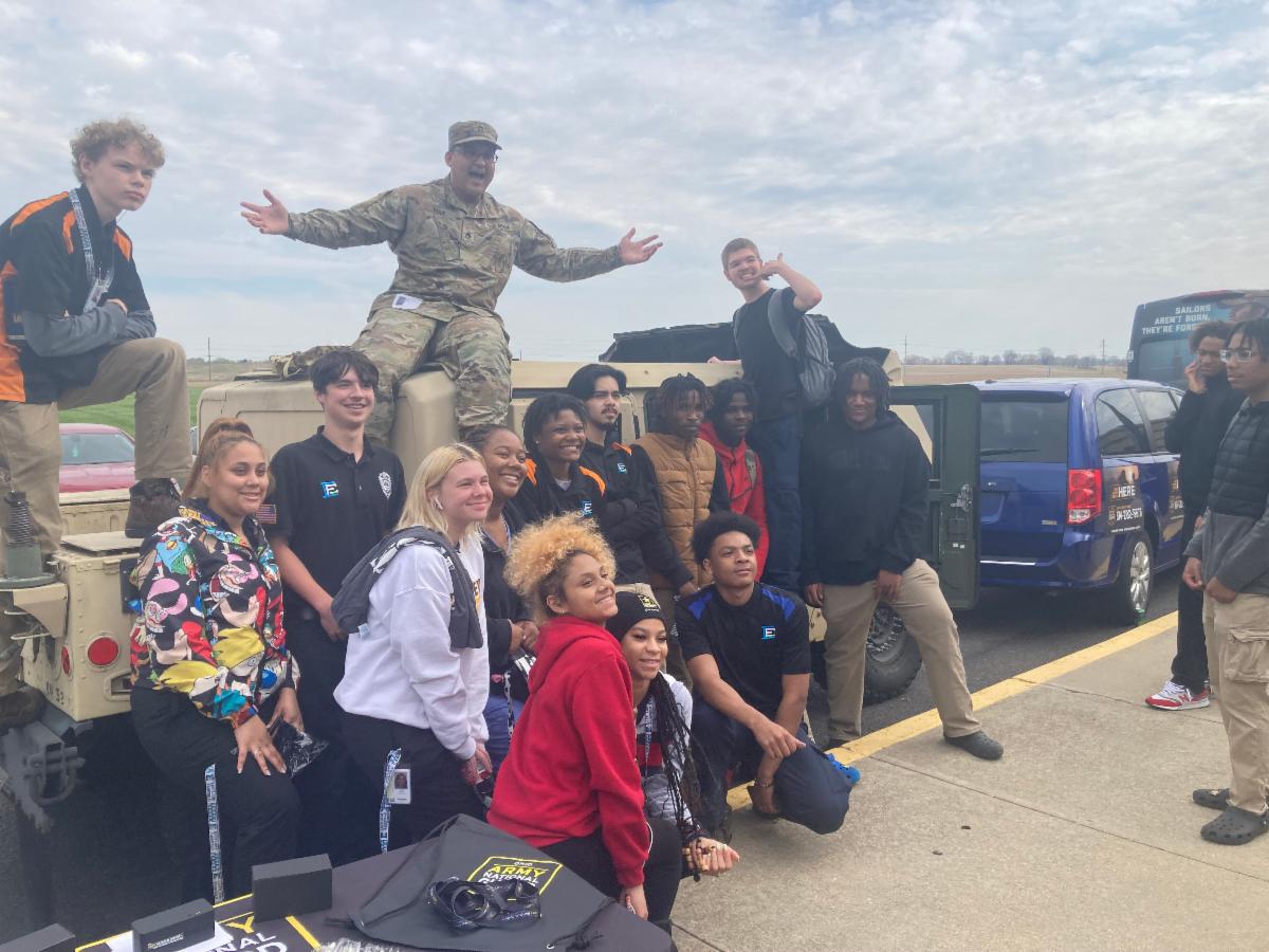Members of the military and students gather around a military vehicle.