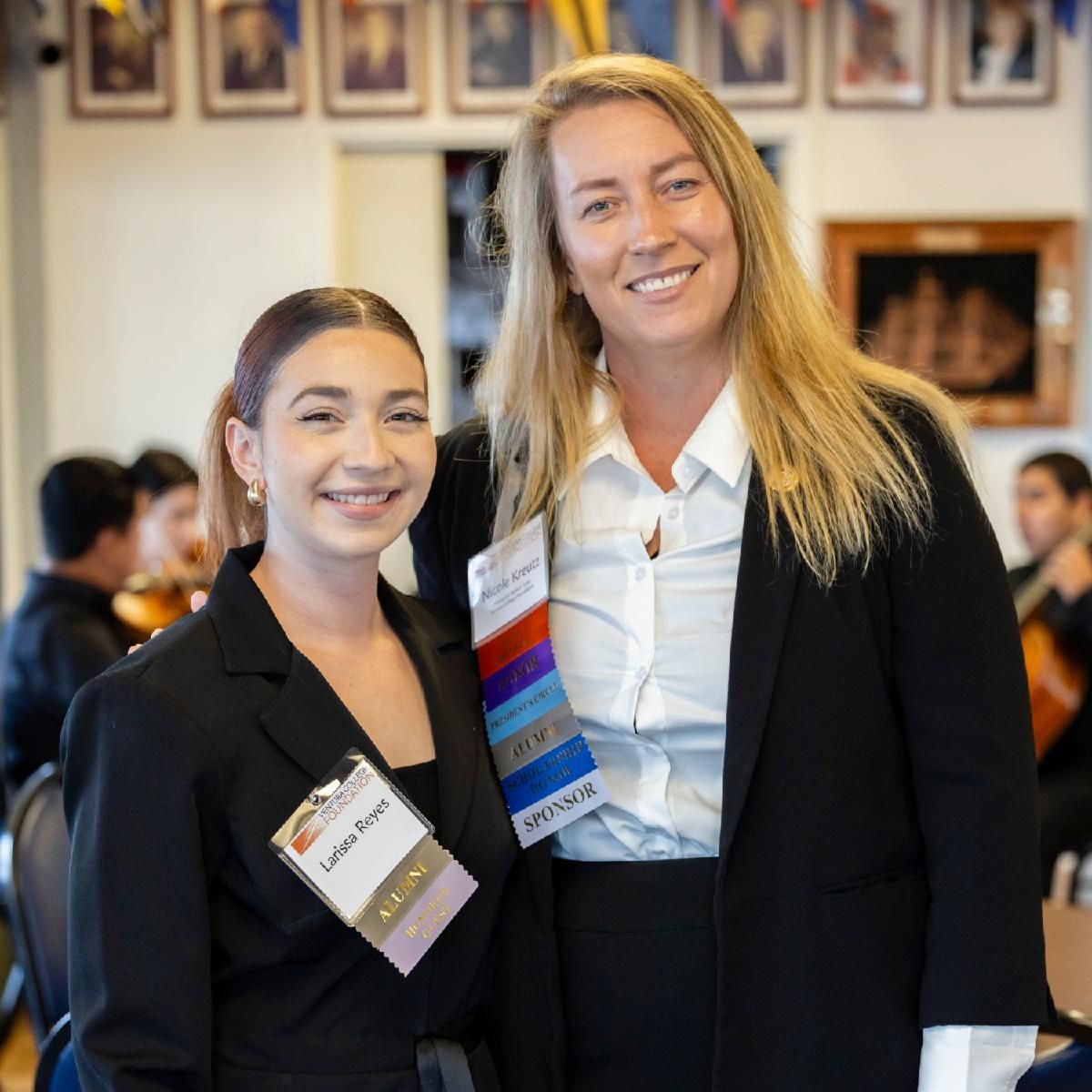 Two women stand side by side smiling at the camera during an indoor gathering. Their name tags read “Larissa Reyes” and “Nicole Kreutz.” Both are dressed in business attire with event ribbons attached to their badges