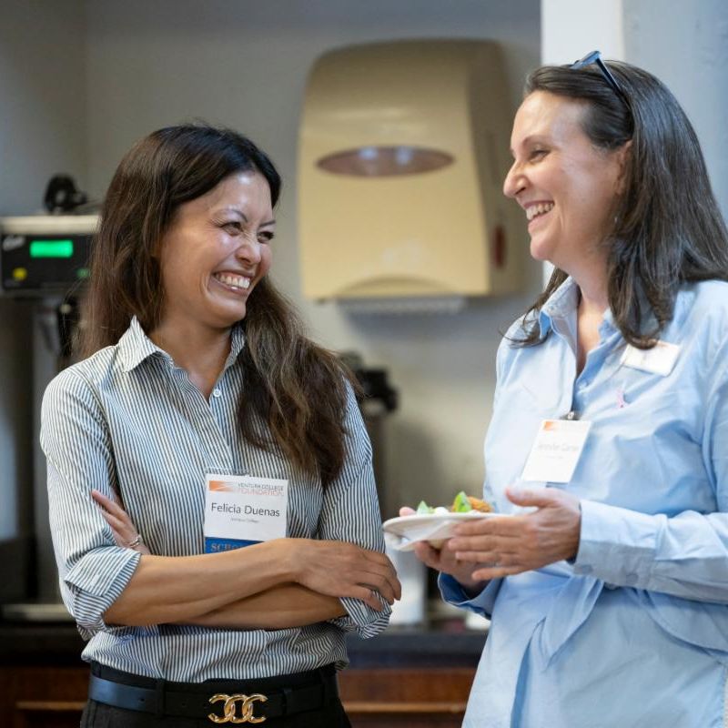 Two women are chatting and laughing together at a social event One holds a plate of food and their name tags read Felicia Duenas and Jennifer Garner They are standing indoors near a counter and kitchen equipment.