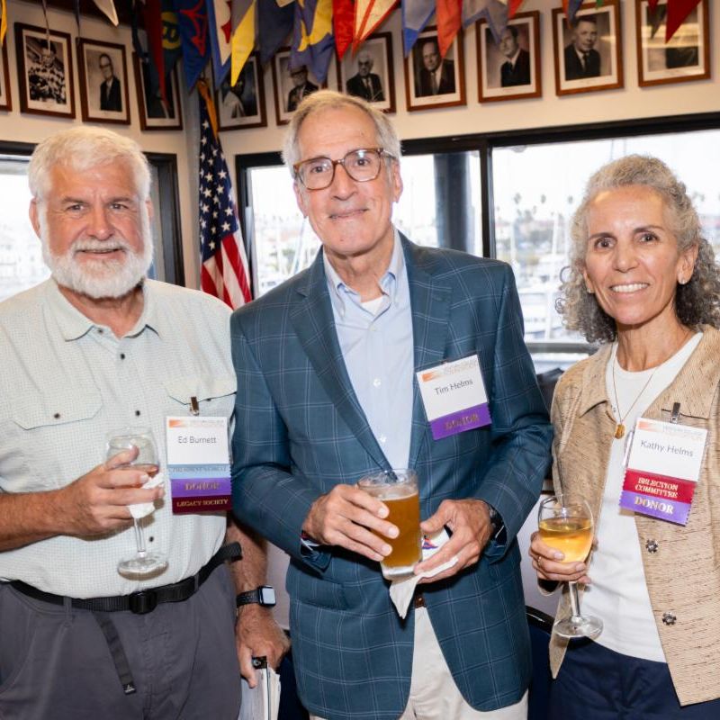 Three people stand together smiling at an indoor event. They each hold a drink and wear name tags that read “Ed BurnettTim Helms and Kathy Helms  Behind them are windows showing a marina and framed portraits on the wall