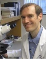 Brunette man in lab coat smiling next to a microscope