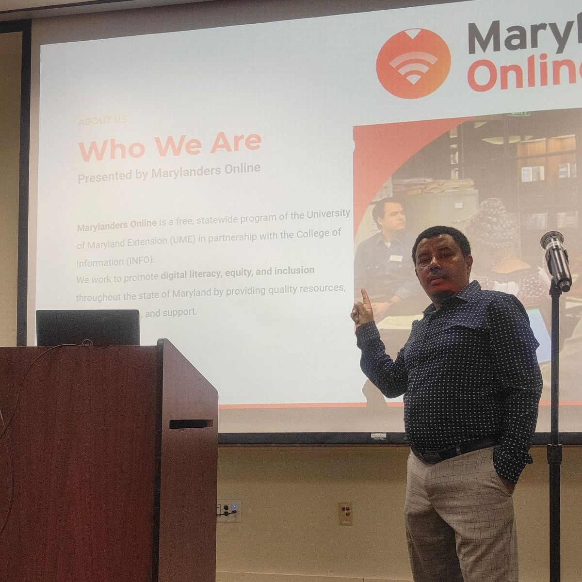 Picture of UME Tech Educator standing next to a podium pointing at a projector screen behind him that shares information about Marylanders Online