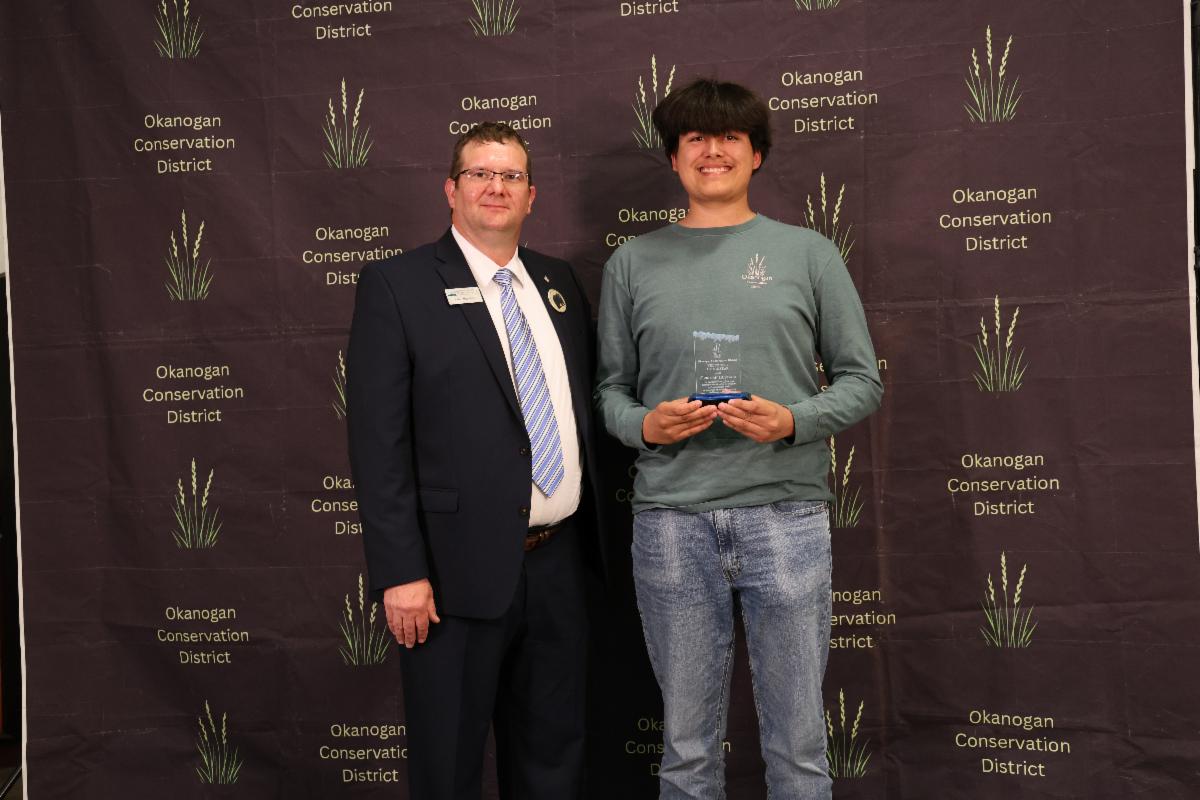 Volunteer, Cristian Hurtado and NRCS Earth Team State Coordinator, Kris Mills, smiling and holding Volunteer of the Year award in front of backdrop.