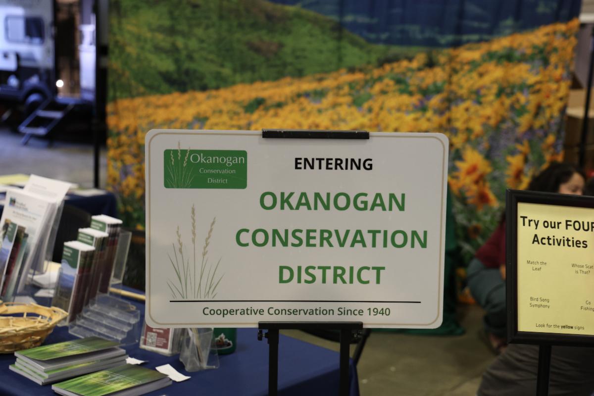 A sign with the words, Entering Okanogan Conservation District is in the foreground. A table with flyers and banner with flowers can be seen in the background.