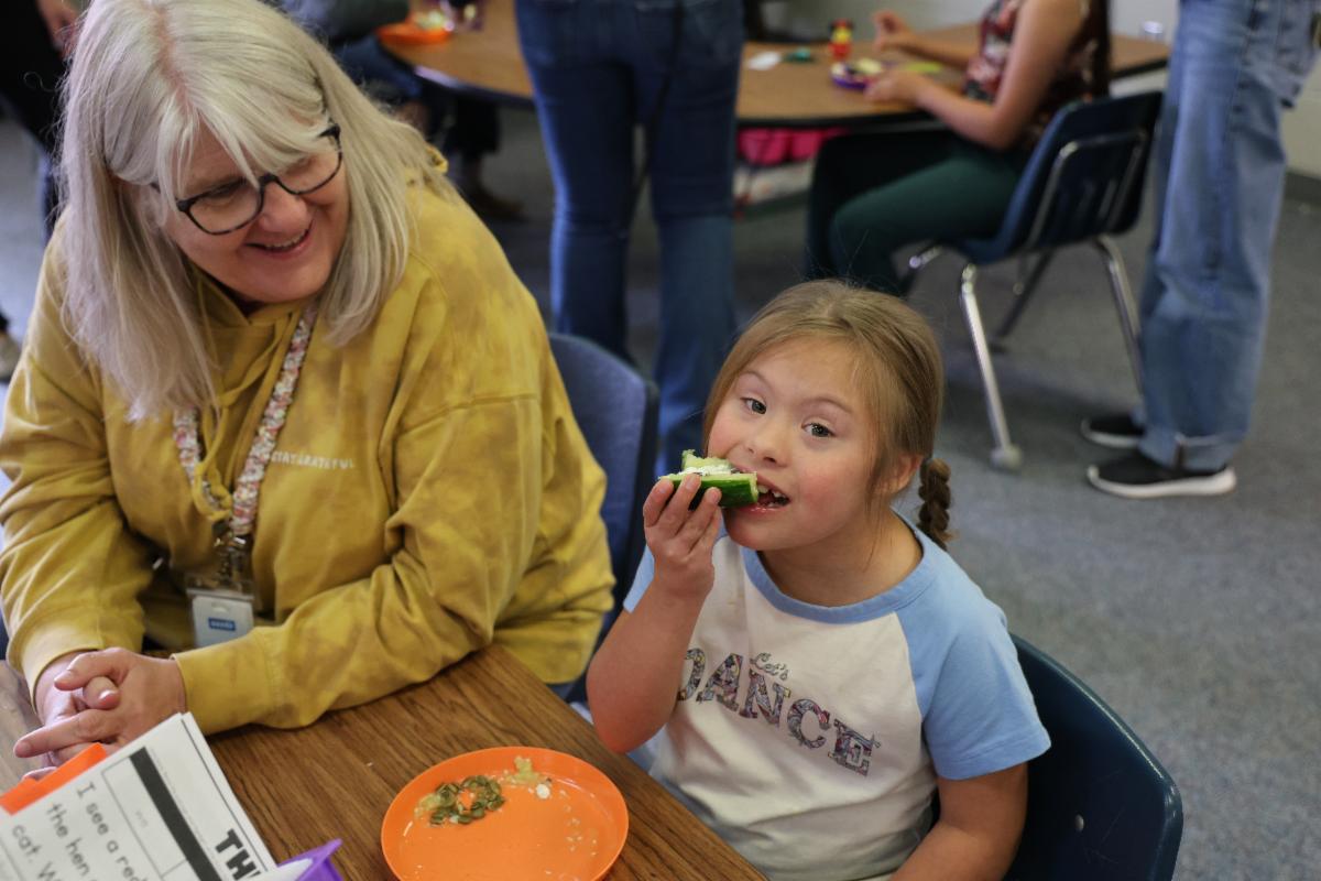 A girl and an adult sit at a table in a classroom, the girl is eating a cucumber