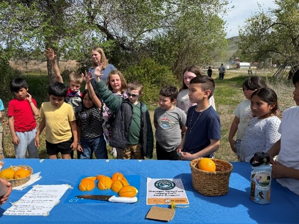 10+ students smiling around a table with oranges on the table.