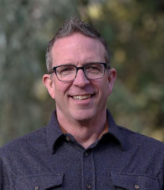 Executive Director, Craig Nelson, smiling and wearing a black dress shirt with jeans, trees in background