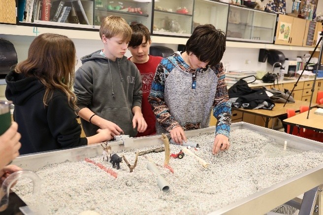 a few students gathering around the stream table, hands in the sand and rearranging a farm model.