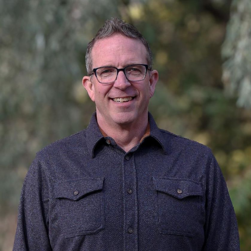 Executive Director, Craig Nelson, smiling and wearing a black dress shirt with jeans, trees in background