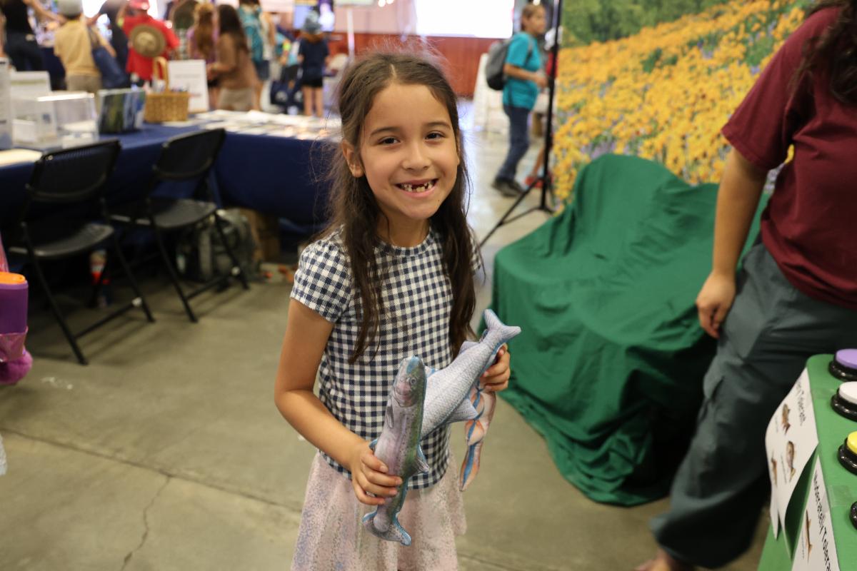 A girl smiles and holds a stuffed fish. In the background is a table with flyers and a banner with yellow flowers.