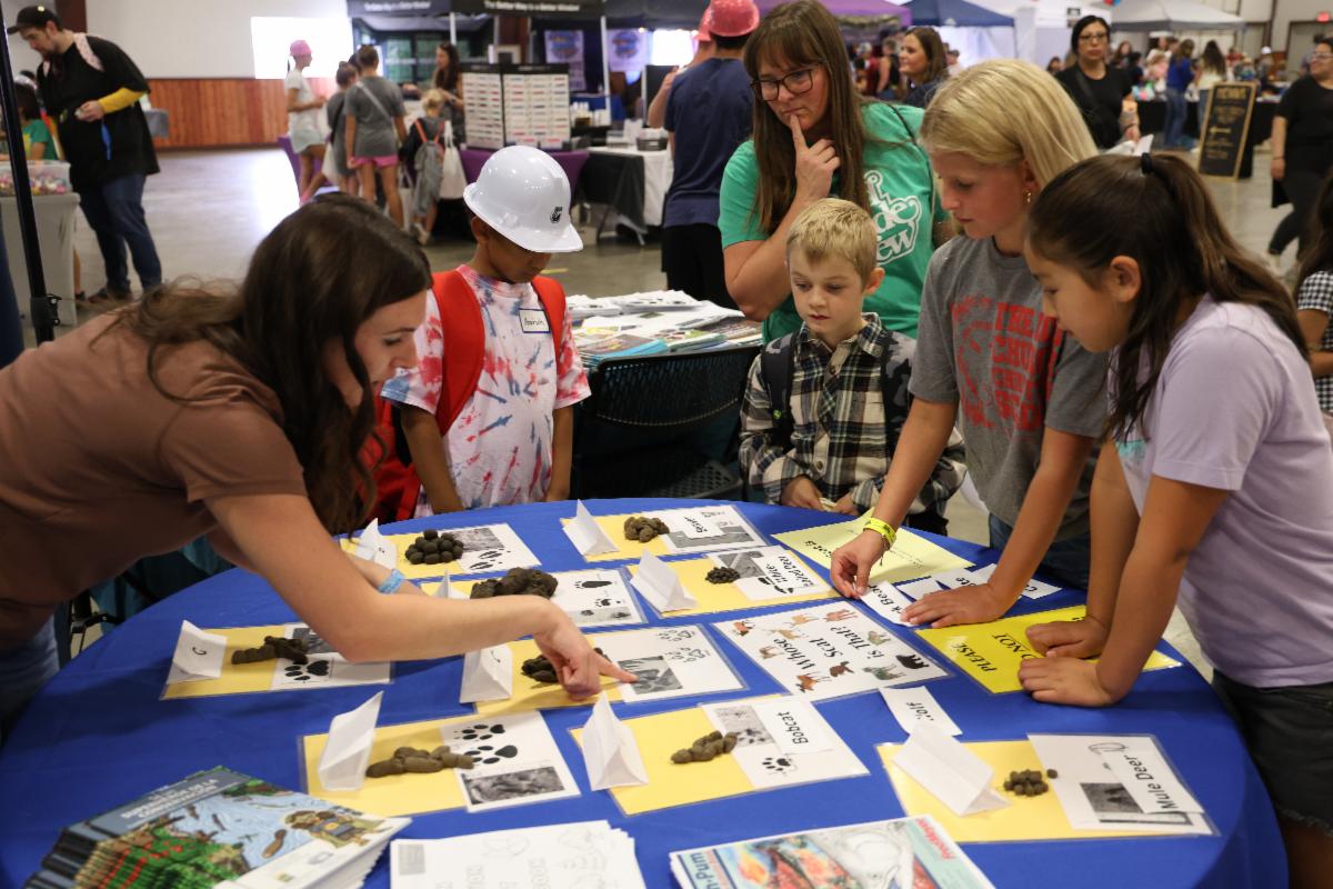 Four children stand around a table with plastic animal scat and photos of animal footprints. A staff member points to one of the footprints.