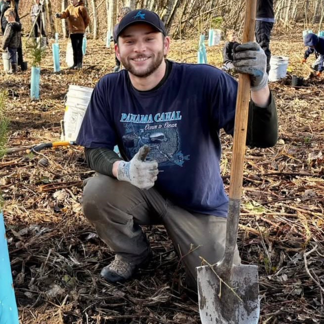Summer intern, Jack Earnest, smiling, kneeling, and holding a thumbs up with a shovel at a planting project.