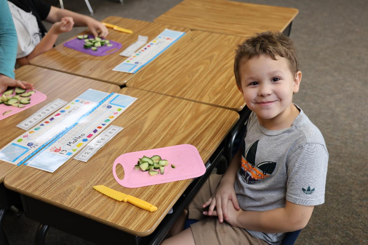 A child sits and smiles next to a cutting board with cut up cucumber.