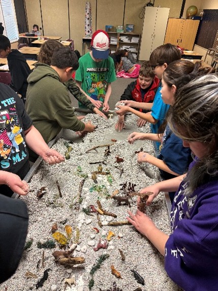 ten students gathered around the stream table with hands in the sand with various items in the sand.