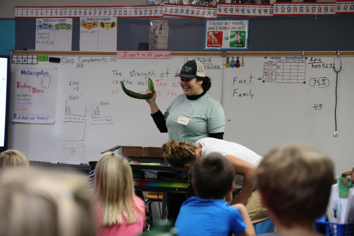 Students watch, Emily Bustamante, as she smiles and holds up a cucumber 