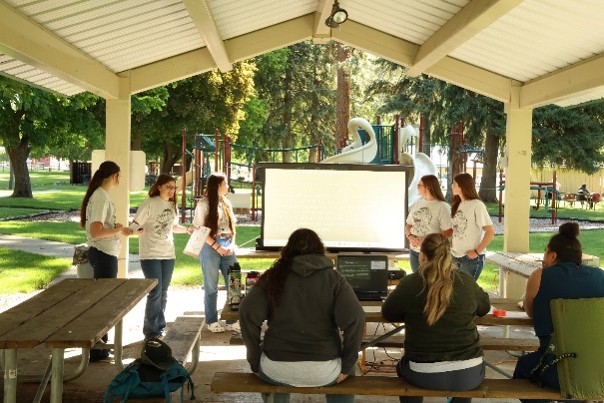 Students under a covered area in front of a screen giving a presentation.