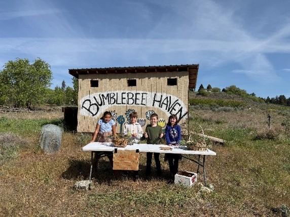 Four students from the school garden pictured at a table smiling with a shed in the background labeled "Bumble Bee Haven" 