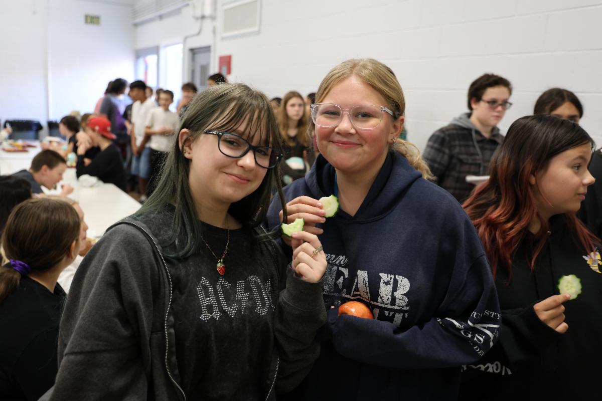 Two middle schoolers hold up cucumber slices in the lunch room and smile.