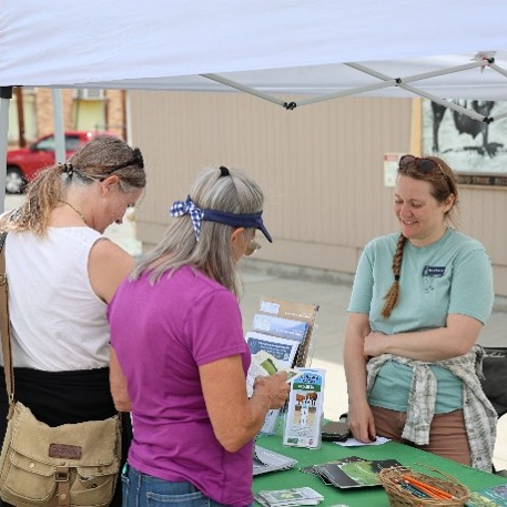 Two adults talk to staff member Chevelle Yeckel. Chevelle is smiling and standing at a table with flyers. 