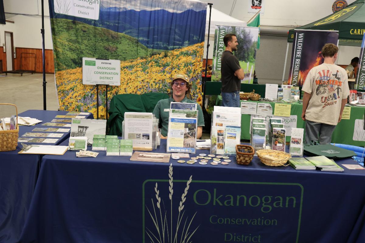 Staff member, Eli Loftis, sits smiling at a table with flyers. Behind him there is a banner with flowers.