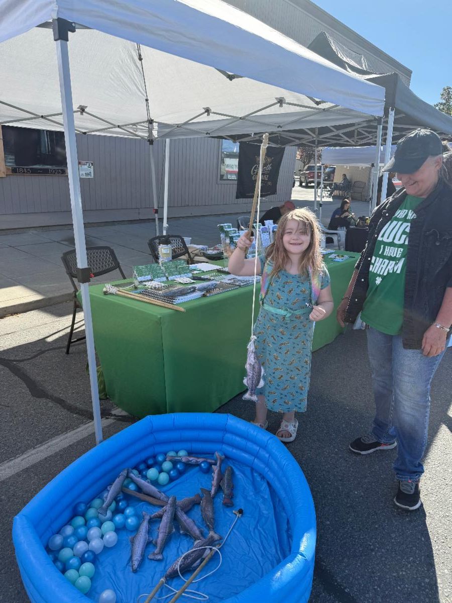 A girl stands behind a blow-up pool and holds up a fishing poll with a magnetic fish on the end. She is smiling. There is a table behind her with flyers.. 