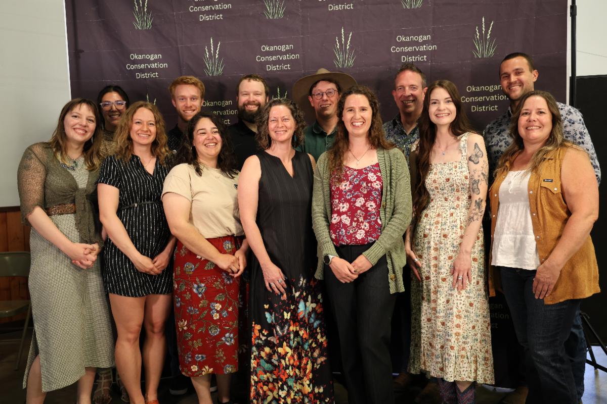 13 staff members in business casual attire, smiling for a group photo at the Conservation Celebration event in front of backdrop with "Okanogan CD" repeating text.