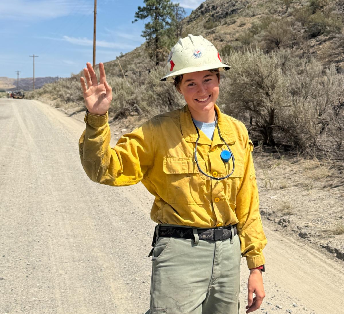 New staff member, Rowan, smiling and waving to the camera, wearing yellow wildfire protective gear and hard hat, standing on a dirt road.