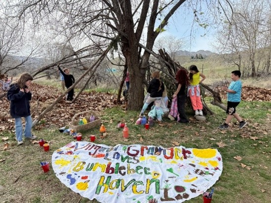 Kids playing in a stick fort at the school garden with a banner on the ground that says "Virginia Grainger loves Bumble Bee Haven."