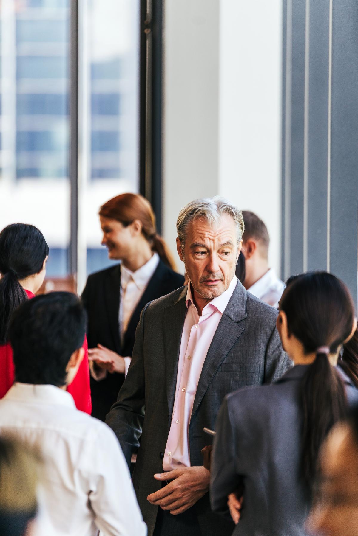 Professionals in suits network and converse at a corporate event near large windows in a modern office building.