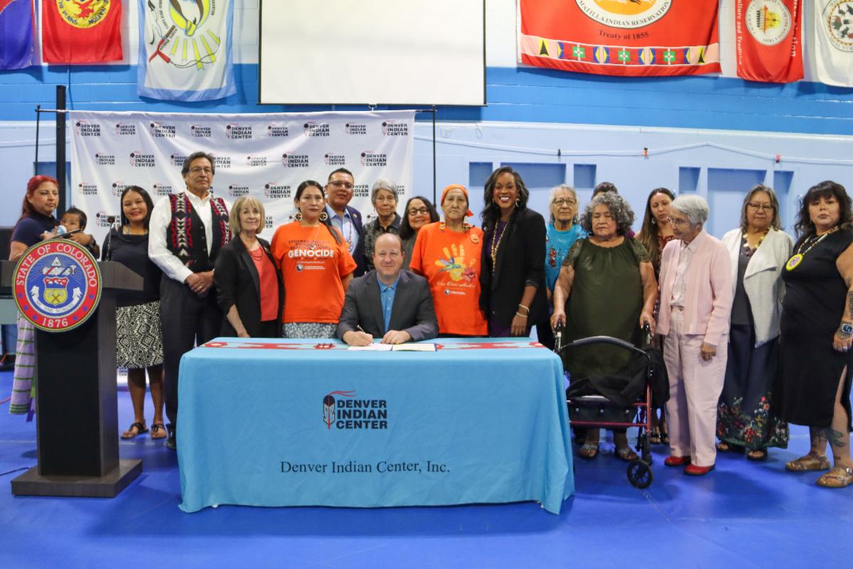 Governor Polis signs HB24-1444 at the Denver Indian Center at a table surrounded by advocates and legislators from the Native American community.